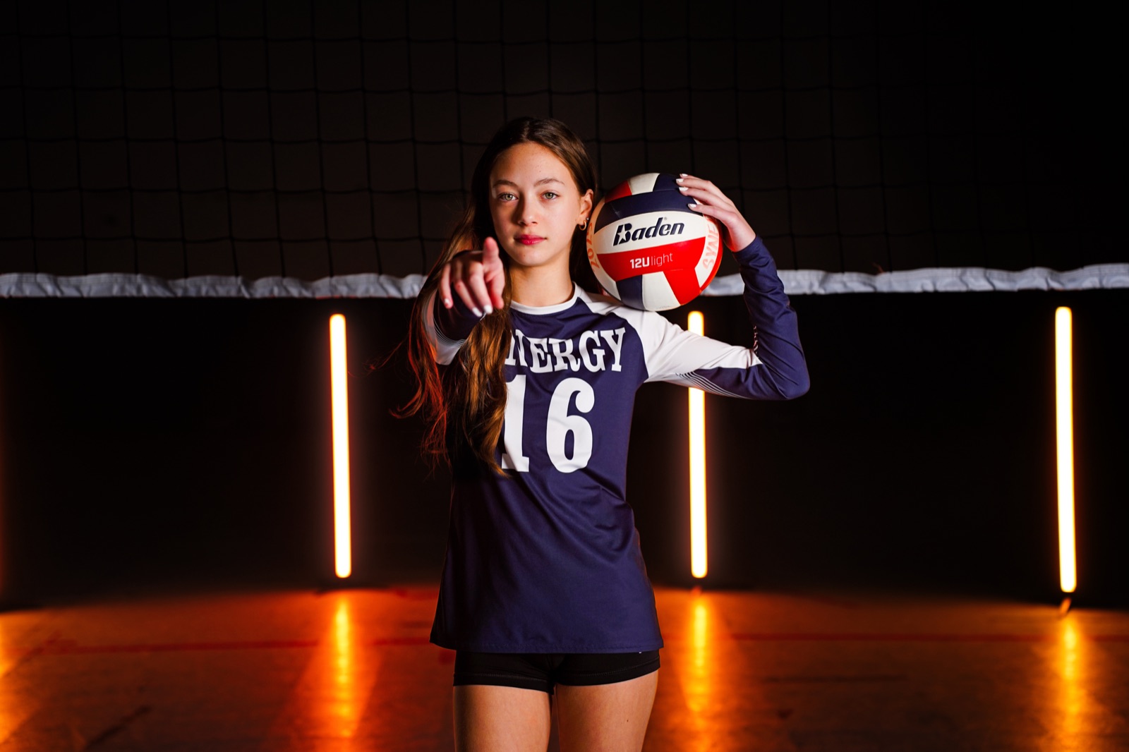 Volleyball player pointing at the camera with a ball on her shoulder, lit by orange light tubes